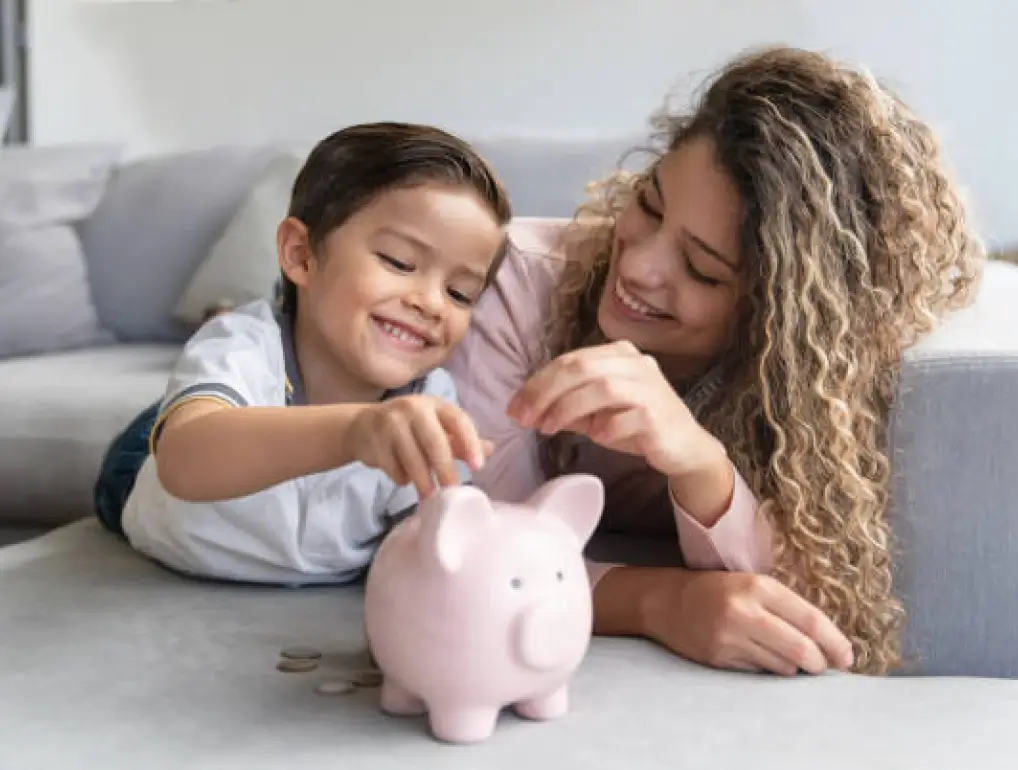 Mother and son putting money in oink piggy bank and smiling together on grey couch